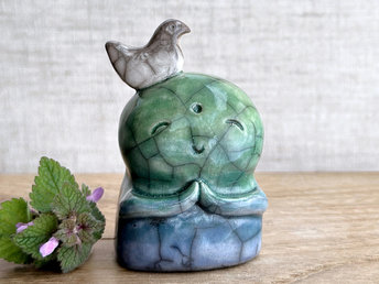 rweb patterns in the glaze, in addition to the normal raku crackles, from where raindrops fell on it.a raku ceramic jizo nature spirit guardian sculpture glazed in a bottom-to-top gradient of blue to forest green to spring green. they have a happy, smiley face, little praying hands, and a sweet white bird on their head. there are tiny dark grey spide