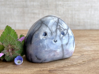 small cloud raku ceramic sculpture glazed in a bottom-to-top gradient of violet to blue to pale blue, almost white at the top. it has an adorable, happy face and is shaped vaguely like a squat raindrop. there are tiny dark grey spiderweb patterns in the glaze, in addition to the normal raku crackles, from where raindrops fell on it.