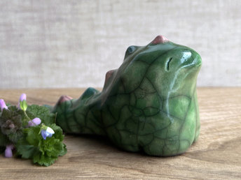 raku ceramic chunky dragon sculpture. it's glazed in green and has a very friendly face. it also has a long tail with little triangle bumps that run from the top of its head to the base of its tail; they're glazed in alternating forest green and soft red pink. 