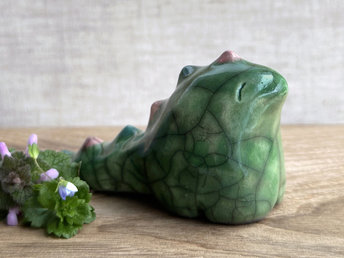 raku ceramic chunky dragon sculpture. it's glazed in green and has a very friendly face. it also has a long tail with little triangle bumps that run from the top of its head to the base of its tail; they're glazed in alternating forest green and soft red pink. 