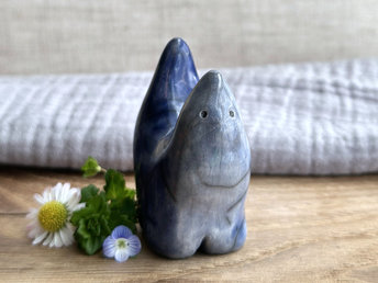 raku ceramic creature glazed in a front-to-back gradient of pale blue-grey to a darker, more intense blue colour. it has four stubby legs and both its front and back ends rise up to a point, so that when you look at it from the side, it almost looks like a pair of horns. it has a small, cute face.