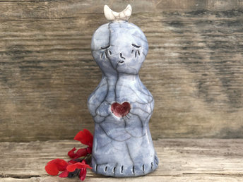 Tall raku ceramic sculpture of a smiling being. She is glazed is soft blue and has a white crescent moon on her head. In her chest is a cut-out heart shape glazed in warm soft red.