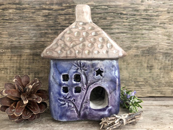 Raku ceramic kurinuki cottage glazed in soft indigo and violet with carved flower and tree patterns on the walls. The roof has a chimney, patterning, and is glazed in pale brown. This house has a cute little doorway and lots of windows.