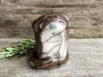 Adorable river otter raku sculpture glazed in brown with a cream-coloured tummy. He has a black nose, little ears, and a tail wrapped around his body. He also has a kind and gentle face.