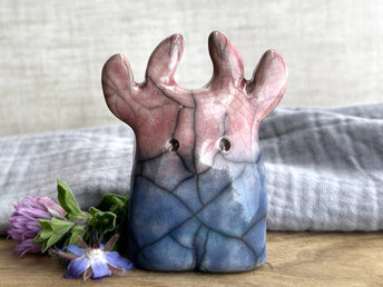 a raku ceramic creature. it has four stubby legs and a wide lower half. it tapers up a little and eventually ends in four small antlers. on the way, it has a very sweet face with a cute little nose. it's glaze in a bottom-to-top gradient of blue to soft reddish pink. it is a friend.