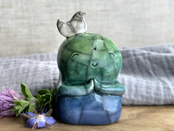 a raku ceramic jizo nature spirit guardian sculpture glazed in a bottom-to-top gradient of blue to forest green to spring green. they have a happy, smiley face, little praying hands, and a sweet white bird on their head.