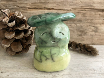 Raku ceramic sculpture glazed in warm, yellow-green to green (bottom to top) with a gentle, smiling face and a darker green leaf on its head.