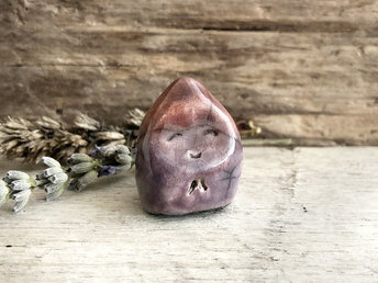 A small, almost (but not quite) teardrop-shaped raki ceramic earth spirit sculpture. It has a gentle, sleepy face and is glazed in soft purple and warm burnt orange colours.