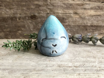 A small raindrop raku ceramic sculpture glazed in soft blue and turquoise colours with a smiley face. A prayer to end the droughts and floods.