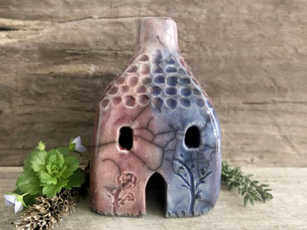 Raku ceramic kurinuki cottage glazed in bordeaux red, warm orange, indigo, and violet. It has a cute little chimney, plant carvings on its walls, a cute door and lots of little windows.