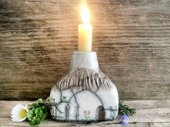 Raku ceramic candlestick in the shape of a rectanglar cottage. The top is brown and carved to look like thatch. The rest is a white cottage with windows, a door, and flower and plant carvings.