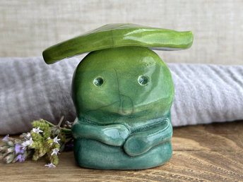 Raku ceramic sculpture glazed in a bottom-to-top gradient of blue to forest green to spring green with a kind, but slightly intense, face, and a spring green leaf on its head. It also has two cute little arms.