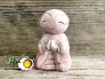 A smiley, loving, happy little Jizo Shinto raku ceramic sculpture in a classic monk form. It is glazed in very pale soft red/pink colour and has a kind, gentle face. Its body is sculpted to look like it's wearing robes, and it has sweet little praying hands.