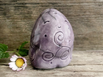 round and vaguely egg-shaped nature spirit sculpture. it is glazed in a soft purple colour and has spiral carvings, including a triskelion, on its body. it also has a gentle, happy face.