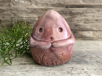 vaguely dome-shaped nature spirit ceramic sculpture. it is glazed a deep-orange / salmon-pink colour and has plant and flower carvings on its body. it also has a gentle face, and two little arms across its front.