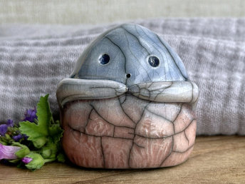 a vaguely dome-shaped nature spirit ceramic sculpture. it is glazed in a bottom-to-top gradient of soft, pale reddish pink to pale blue and has plant and flower carvings on its body. it also has a gentle face, and two little arms across its front.