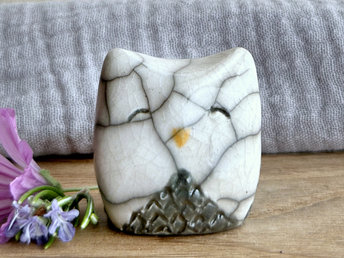Raku ceramic little owl or owlet kami nature spirit statue with sort of pointed ears. It’s glazed in white and grey with gentle, smiling eyes and a little golden yellow beak.