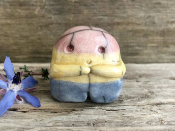 A sweet little nature spirit ceramic guardian statue glazed in the pansexual flag blue, yellow, and pink colours and with a lovely, gentle face and little arms. It has a small branch design carved up one side.