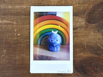 an instax mini print of a blue kodama spirit with a white crescent moon on their head. there's a wooden stacking rainbow behind them and they're in a shaft of light.