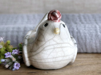 a white raku ceramic chicken with grey eyes, a pale orange beak, and a bordeaux red comb on the top of her head. she looks very sweet.