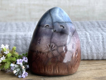 vaguely dome-shaped nature spirit ceramic sculpture. it is glazed in a bottom-to-top gradient of a brown to soft mauve to violet to blue and has autumnal plant and flower carvings on its body. it has a gentle, smiling face.