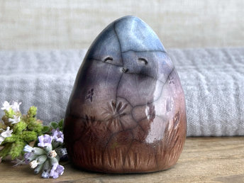 vaguely dome-shaped nature spirit ceramic sculpture. it is glazed in a bottom-to-top gradient of a brown to soft mauve to violet to blue and has autumnal plant and flower carvings on its body. it has a gentle, smiling face.