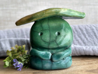 Raku ceramic sculpture glazed in a bottom-to-top gradient of forest green to leaf green with a kind, but slightly intense and worried, face, and a leaf-green and golden-yellow leaf on its head. It also has two cute little arms.