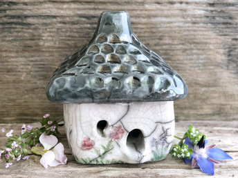 raku ceramic kurinuki cottage glazed in white with flowers and tree patterns carved on the walls and glazed in colour. the roof has a chimney and is carved and glazed to look like grey stone roof tiles. the cottage has a cute little doorway and lots of windows.