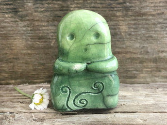 A sweet kodama nature spirit ceramic guardian statue glazed in shades of green and with a lovely, gentle face and little arms. it has celtic spiral designs carved on its front.