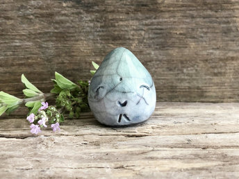 A small raindrop raku ceramic sculpture glazed in a gradient of pale blue and aqua green (bottom to top) with a smiley face. A prayer to end the droughts and floods.