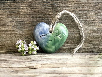 A smiley blue and green heart ornament in raku ceramic. It has a hole in the top, and there is a piece of twine threaded through the hole to act as a hanger.