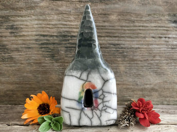Raku ceramic spirit house in the shape of a church. The top is a steeple, grey and carved to look like slates. The rest is a white square building with a door and several windows, and each window has glaze around it in lots of colours to emulate light from stained glass.