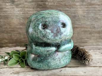 A kodama nature spirit ceramic guardian statue glazed in a turquoise and copper glaze. It has a wide face, two big eyes, and cute little arms.