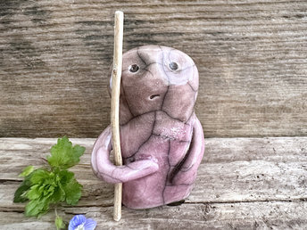 raku ceramic nature spirit warrior sculpture. it is glazed in a gradient from mauve to lighter soft mauve-brown (bottom to top) and carries a brown stick in one of its arms. it has a concerned but determined look on its gentle face.