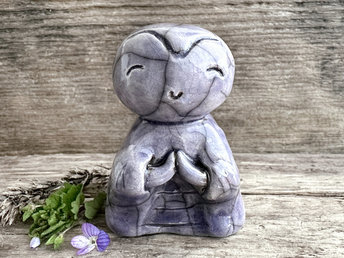 A smiley, loving, happy little Jizo Shinto raku ceramic sculpture in a classic monk form. It is glazed in a soft violet colour and has a kind, gentle face. Its body is sculpted to look like it's wearing robes, and it has sweet little praying hands.