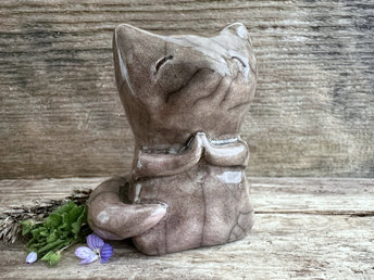 Raku ceramic fox sculpture with a kind, gentle face and sweet praying hands. It is glazed in a warm brown colour with a hint of soft pink, and its tail is curled around its body.