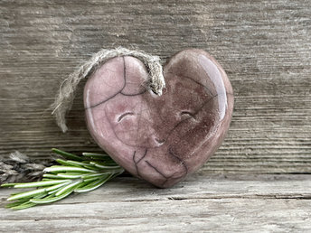 A smiley heart ornament in raku ceramic and glazed in soft light brown and mauve. It has a hole in the top, and there is a piece of twine threaded through the hole to act as a hanger.