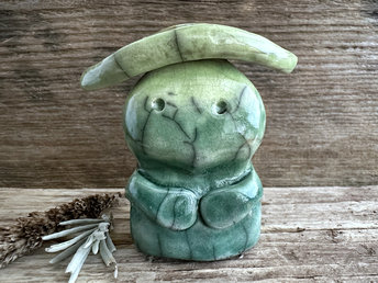 Raku ceramic sculpture glazed in forest green to spring green (bottom to top) with a sweet face and a lighter, yellow-green leaf on its head. It also has two cute little arms.