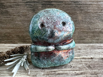 A kodama nature spirit ceramic guardian statue glazed in a turquoise and copper glaze. It has a wide face, two big eyes, and cute little arms.