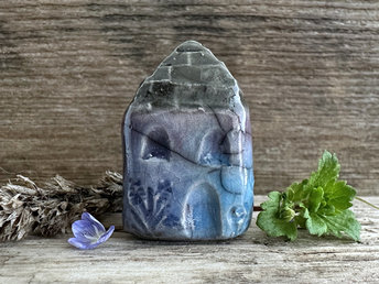 Tiny raku ceramic cottage glazed in blue, violet, and turquoise. Its roof is glazed in grey and carved to look like slates. It has a carved door and windows and lots of plant and flower carvings too.