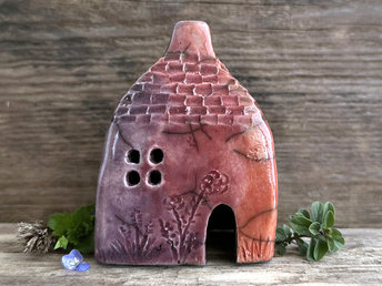 Raku ceramic kurinuki cottage glazed in bordeaux red, warm orange, mauve, and purple. It has a cute little chimney, plant carvings on its walls, a cute door and lots of little windows. Its roof is carved to look like slates.