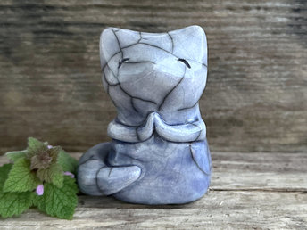 Raku ceramic fox sculpture with a kind, gentle face and sweet praying hands. It is glazed in a bottom-to-top gradient of blue to lighter blue, along with a hint of mauve. Its tail is curled around its body.