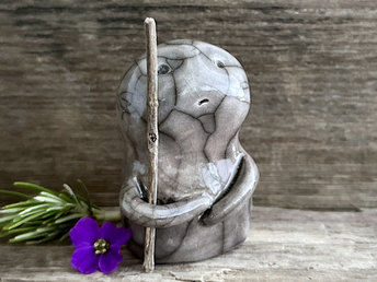 raku ceramic nature spirit warrior sculpture. it is glazed in a gradient from darker to lighter soft grey-brown (bottom to top) and carries a brown stick in one of its arms. it has a concerned but determined look on its gentle face.