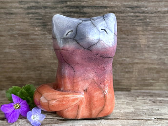 Raku ceramic sitting fox glazed in a gradient from burnt orange to mauve to indigo blue (bottom to top). It has a kind face, and its tail is curled around its body.