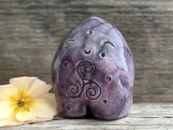 round and vaguely egg-shaped nature spirit sculpture. it is glazed in a mix of purple and violet colours and has spiral carvings, including a triskelion, on its body. it also has a gentle, happy face.