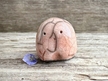 a small and stout nature spirit raku sculpture glazed in a peach colour. it has a cute little face, and a little plant carving on its right side.