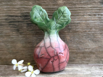a very smiley and happy radish spirit raku ceramic sculpture. it has a round body glazed in a soft radish red colour. the top of its body fades to white, and it has two green leaves on the top of its head. body. it is round with green leaves on top!