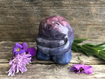 Raku ceramic hug spirit glazed in blue, violet, and purple. He looks very kind and loving and has big arms with which he's hugging himself. His face is gentle and smiling.