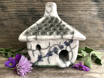 Raku ceramic kurinuki cottage glazed in white with green and purple lavender flower patterns on the walls. The roof has a chimney and is glazed in white, pale green, and light brown to look like thatch. It has a cute little doorway and lots of windows.