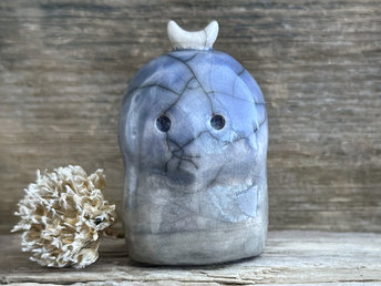 A nature and forest spirit kodama kami ceramic raku sculpture glazed in a bottom-to-top gradient of soft grey-brown to blue. It has a gentle face, and a white crescent moon on its head.